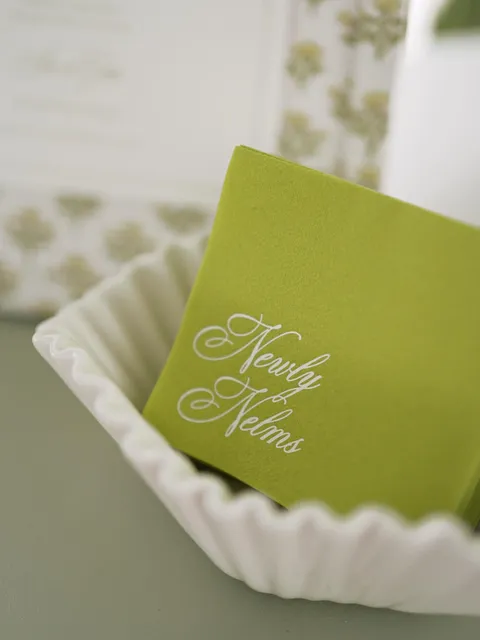Green wedding cocktail napkin by Rachel Latini of Sincerely Rachel with white Juliette calligraphy reading Newly Nelms, styled in a white bowl.