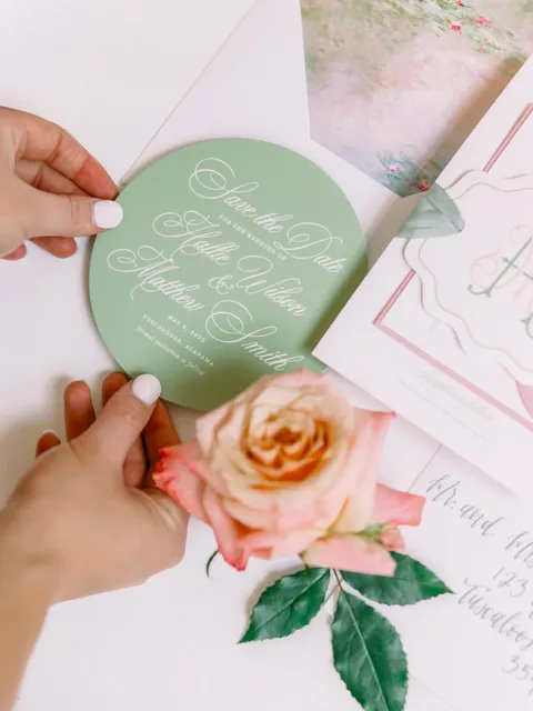 Green oval rehearsal dinner invitation by Lizzie Lettering featuring white Juliette calligraphy, held above pink envelopes and flowers.