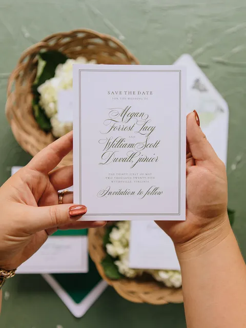 Lavender-bordered save the date card by Anna Howe with Mozart Script calligraphy, held above coordinating wedding stationery.