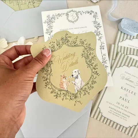 Oval illustrated reception card by Amber LeBlanc Studio featuring Juliette calligraphy, held beside coordinating wedding stationery with botanical borders.