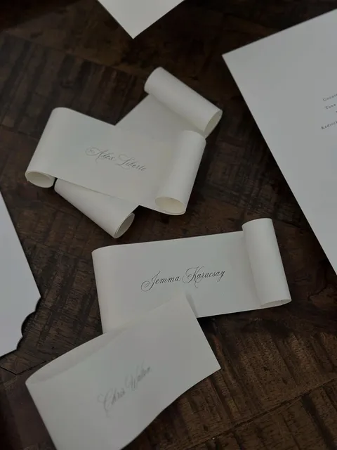 Cream scroll-shaped wedding place cards by Alexandra Collins featuring delicate Juliette calligraphy, arranged on a dark wooden surface.