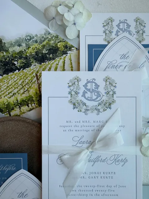 Pale blue Juliette wedding invitation suite by Shop Aurea with a floral crest, blue envelope liner and white ribbon.